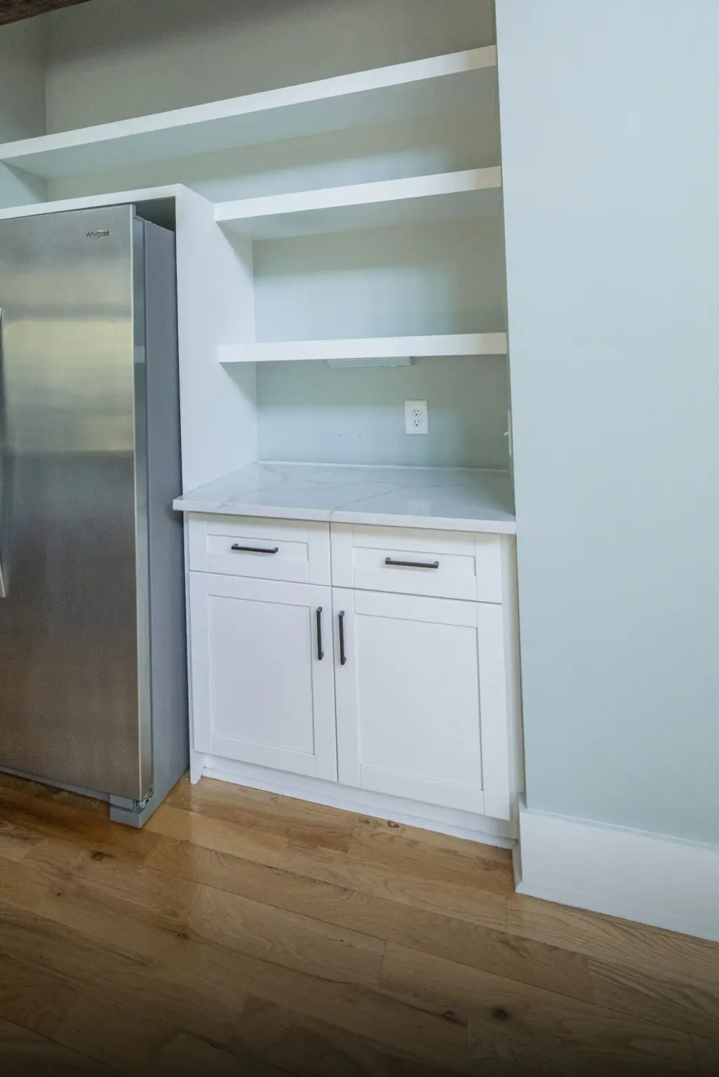 Pantry niche beside a stainless refrigerator with a white veined stone top on a white drawer base and open shelves above