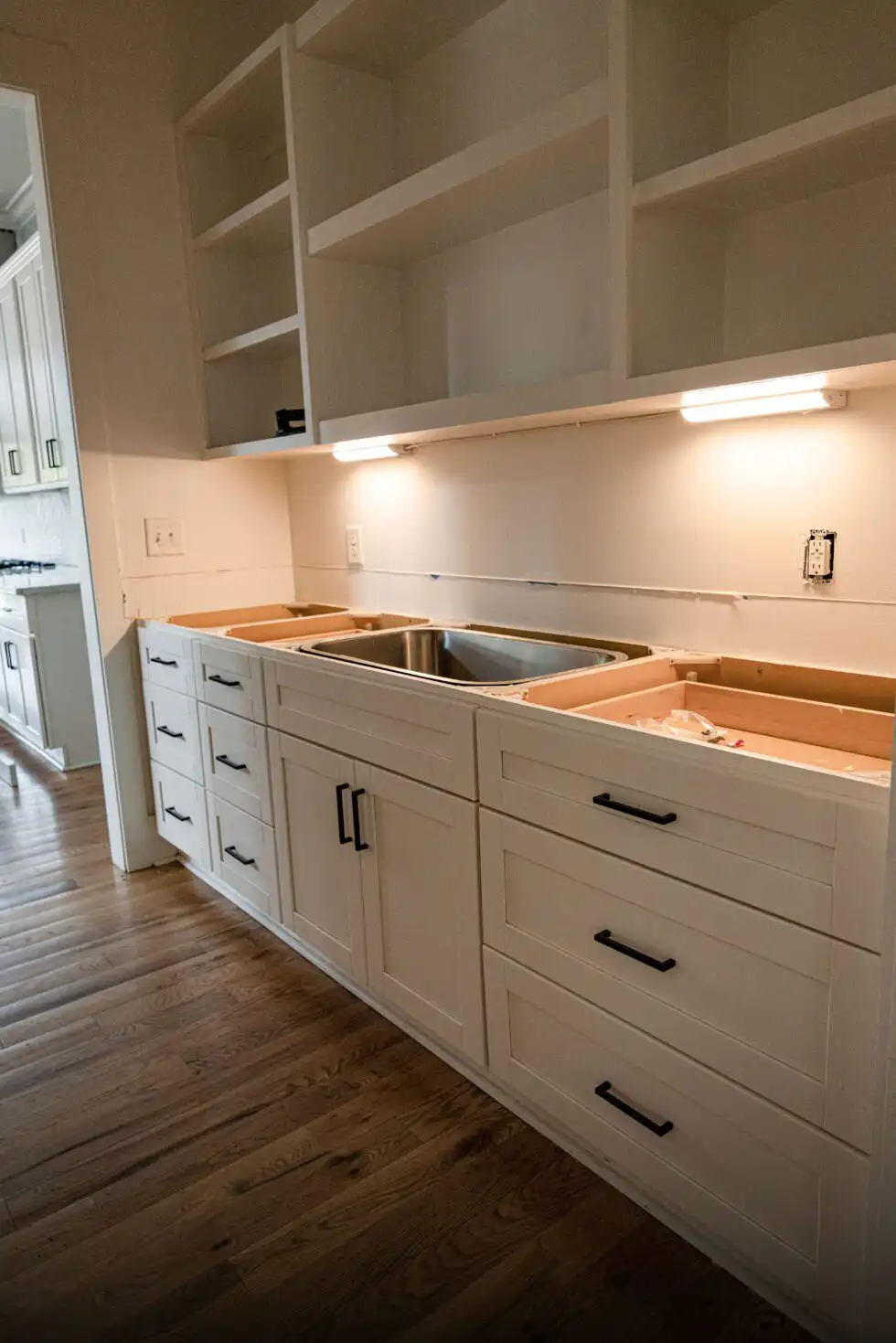 Stainless steel undermount sink resting in a base cabinet run before the stone countertop is installed, with open shelves above