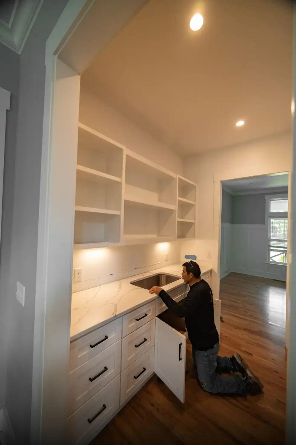 Installer kneeling at an open sink cabinet, checking an undermount stainless sink beneath a white veined stone countertop in a pantry wet bar