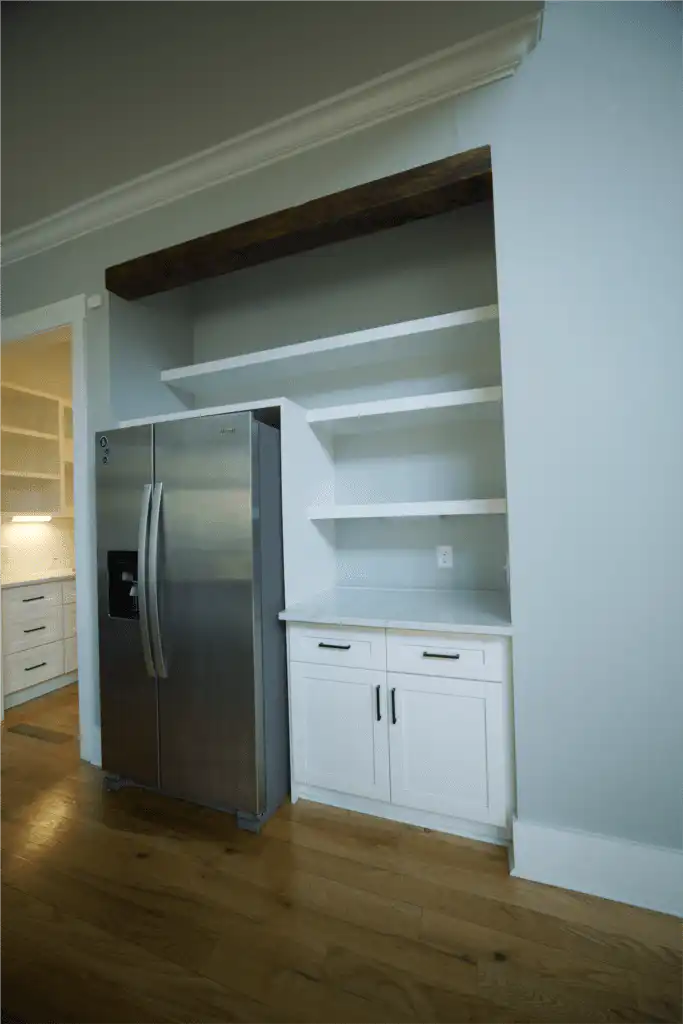 Butler's Pantry Countertops: Plan Them in Seven Decisions Niche beside a side-by-side refrigerator with a white stone countertop on a base cabinet with drawers and doors, open shelves and a wood beam above