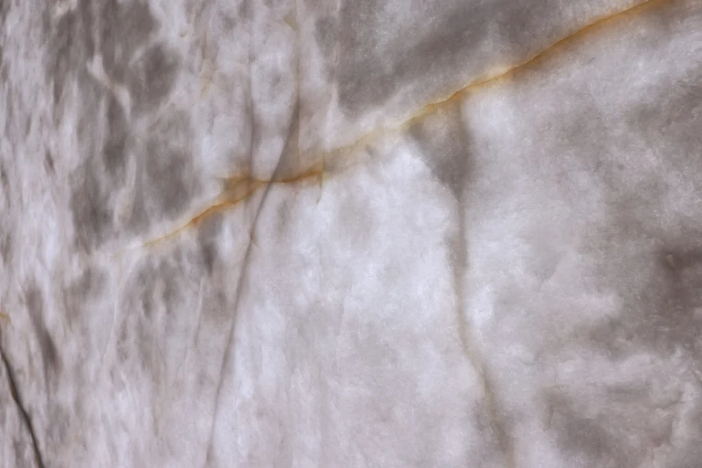 Porcelain vs. Quartzite Countertops: What Each One Is and Where Each Wins Close-up of Crystallo quartzite showing an amber vein and fine gray lines crossing a translucent white body