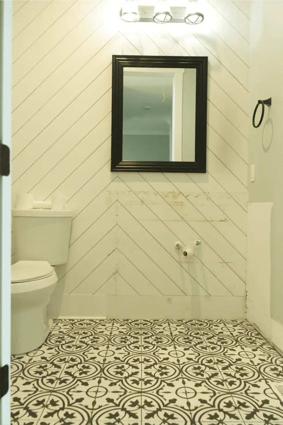 Powder room with a toilet, black-framed mirror, white diagonal shiplap wall and black-and-white patterned floor tile, with no vanity installed yet